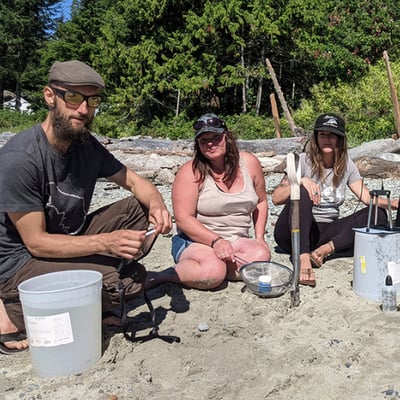 3 community volunteers pose on the beach while conducting a microplastics survey