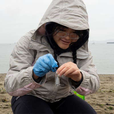 Postsecondary student adds microplastics to a small vial on a rainy beach